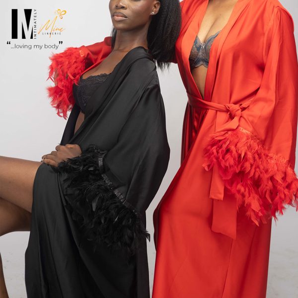 Two women pose in stylish Jamilah robes, one black with feathered trim and the other bright red, showcasing confidence and elegance.