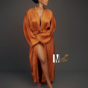 A model stands elegantly in a flowing orange Jamilah satin robe, showcasing a confident pose against a muted backdrop.